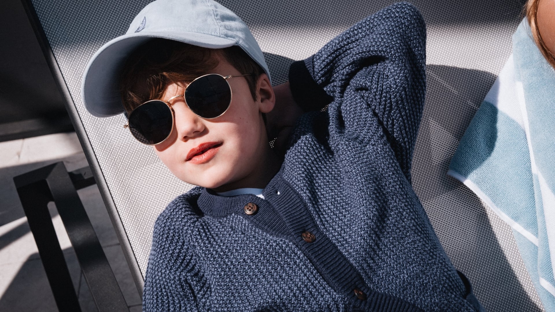 A young boy in sunglasses and a cardigan relaxing on a poolside lounge chair.