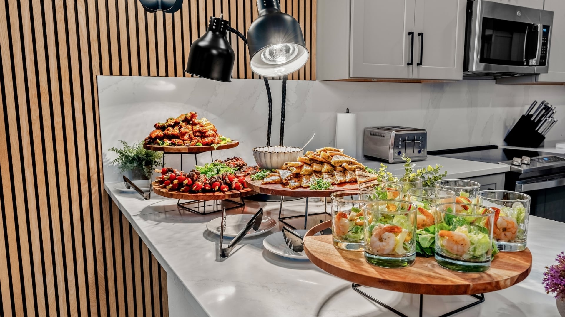 A catering spread on a marble countertop featuring shrimp cocktails, sliders, and chicken skewers under professional heat lamps.